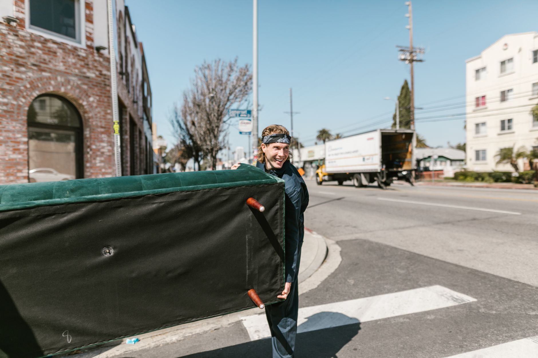 A man carrying a large couch across a sunny street, the same kind of bulky item staged for removal